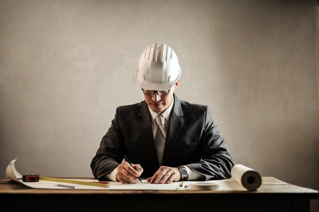 A man wearing a suit and a white hard hat sits at a desk, drawing or writing on architectural plans with a pencil. A measuring tape and rolled-up blueprints are on the table.