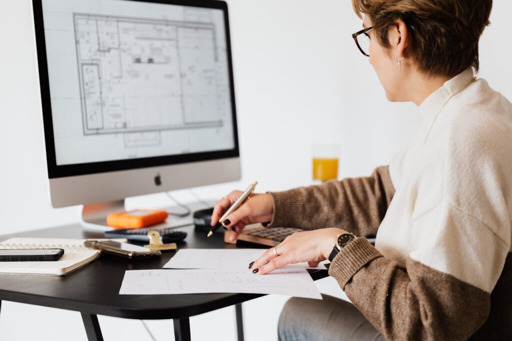 A person sits at a desk working on architectural blueprints, with design plans displayed on a computer monitor and papers, a notebook, phone, and drink nearby.