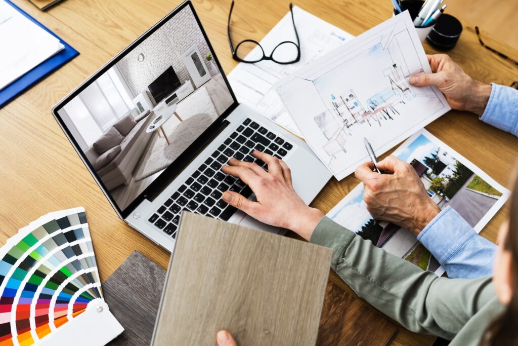 Two people plan an interior design project at a desk, using a laptop displaying a living room, with color swatches, a wood sample, architectural sketches, and glasses spread out on the table.